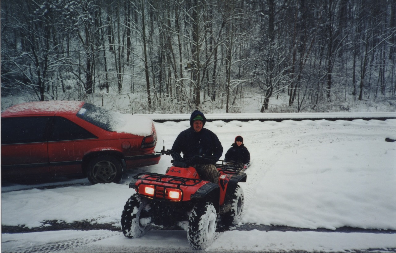 Dad sitting on an ATV with me on a sled in the background. Snow. 