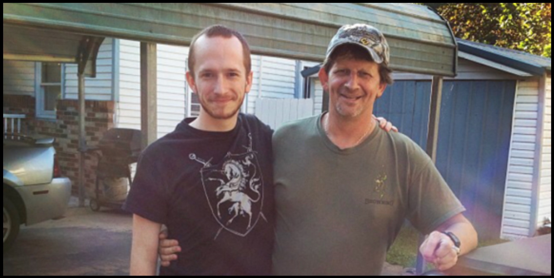 Two men standing together in front of a house. The man on the left has short brown hair and is wearing a black t-shirt with a white design on it. The man on the right has short brown hair and is wearing a green t-shirt and a camouflage baseball cap. They are standing in front of a house with white siding and a blue garage door. There is a silver car parked in the driveway to the left of the men.