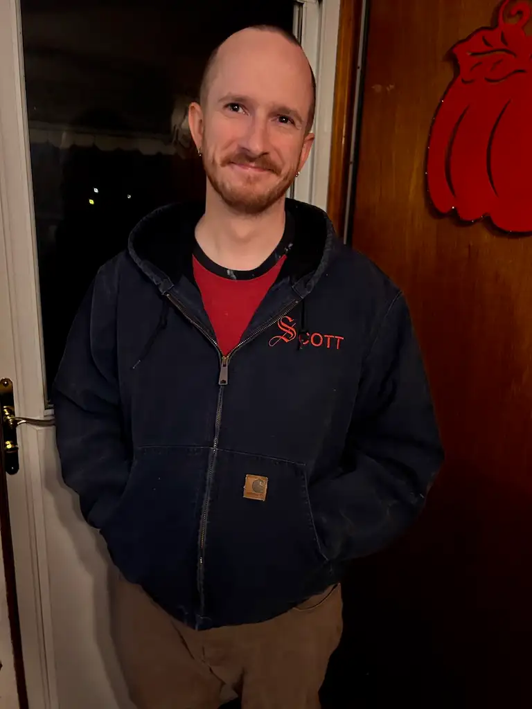 A man standing in front of a door, wearing a dark blue Carhartt zip-up hoodie with 'Scott' embroidered on the chest in red. He has a red shirt underneath and brown pants.