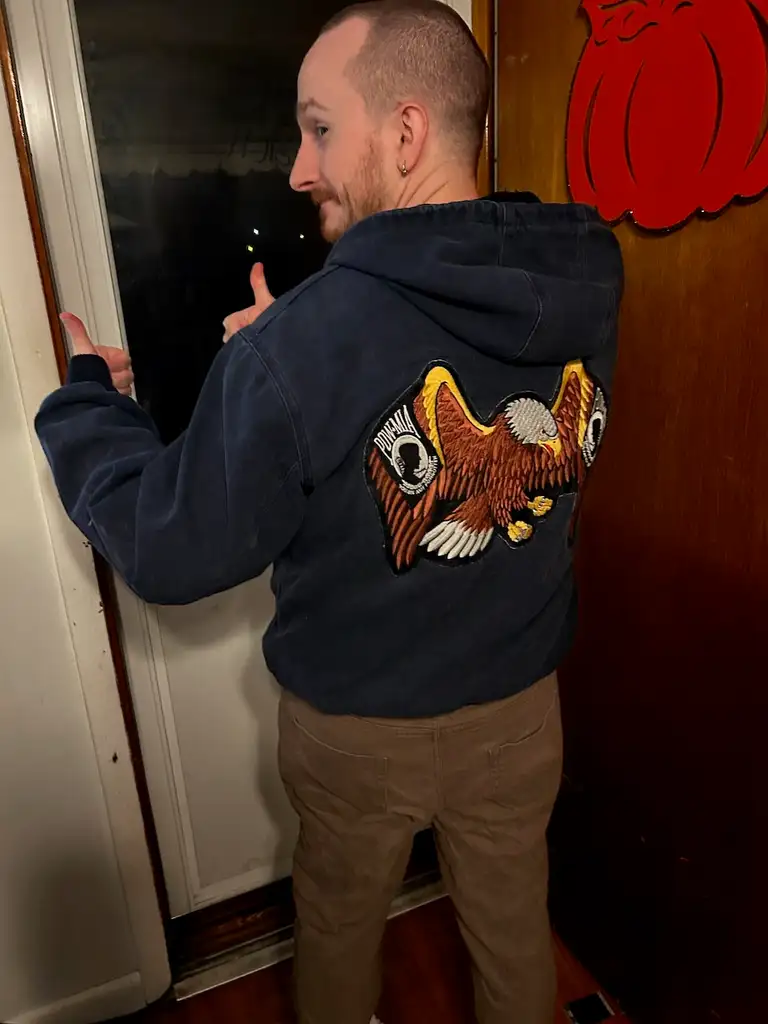 A man standing in front of a door, giving a thumbs-up to the camera. He is wearing a blue hoodie with an eagle design on the back and brown pants.