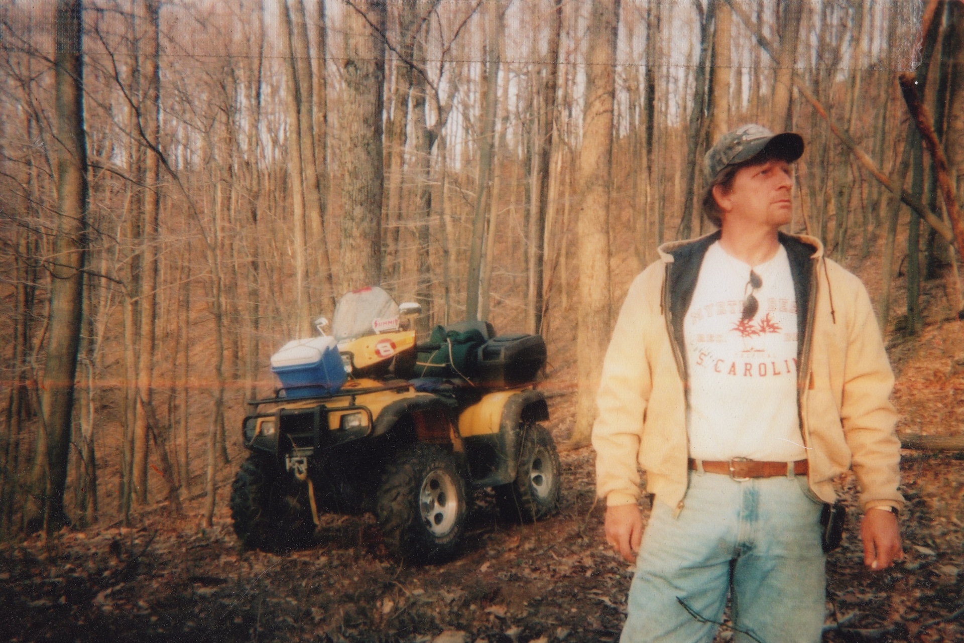 The image depicts a man standing in a wooded area, with an all-terrain vehicle (ATV) parked behind him. The man is wearing a camouflage hat, a tan jacket, and light blue jeans, and is looking to his right. The ATV is yellow and black, with a cooler and other items strapped to the back. The background is a wooded area with tall trees and brown leaves on the ground.