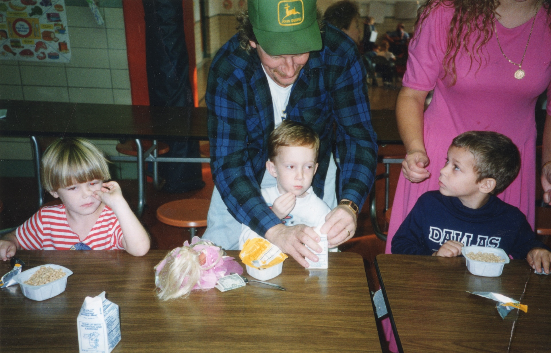 A man helping a young boy eat at a table in a cafeteria. The man is wearing a green baseball cap with a yellow logo and a blue plaid shirt, and is standing behind the boy, who is sitting at a table with a white container of food in front of him. 