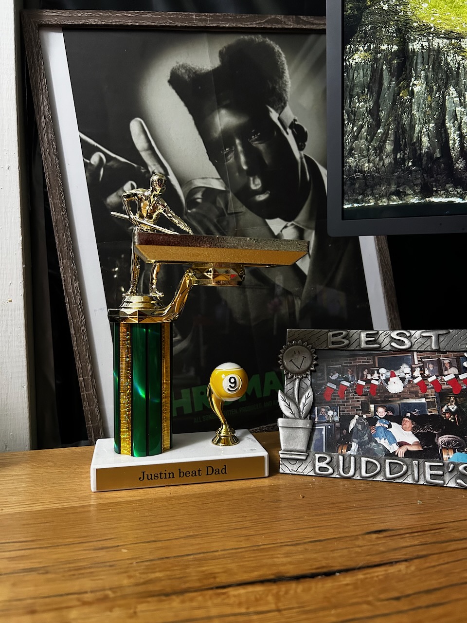 Pool trophy with "Justin beat Dad" plaque sitting on a wooden desk, sandwiched between a framed Tyler, The Creator poster and a framed photo of dad and I that says "Best Buddie's"